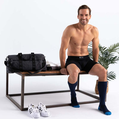 Man sitting on a bench with a duffel bag and sneakers, wearing black Ejis sweat proof boxer briefs and blue socks.