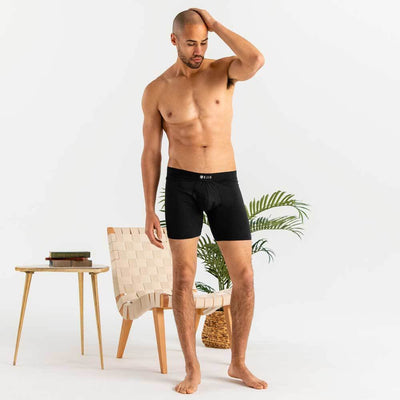 Man wearing black Ejis essential boxer briefs standing in a minimalistic room with a chair and plant.