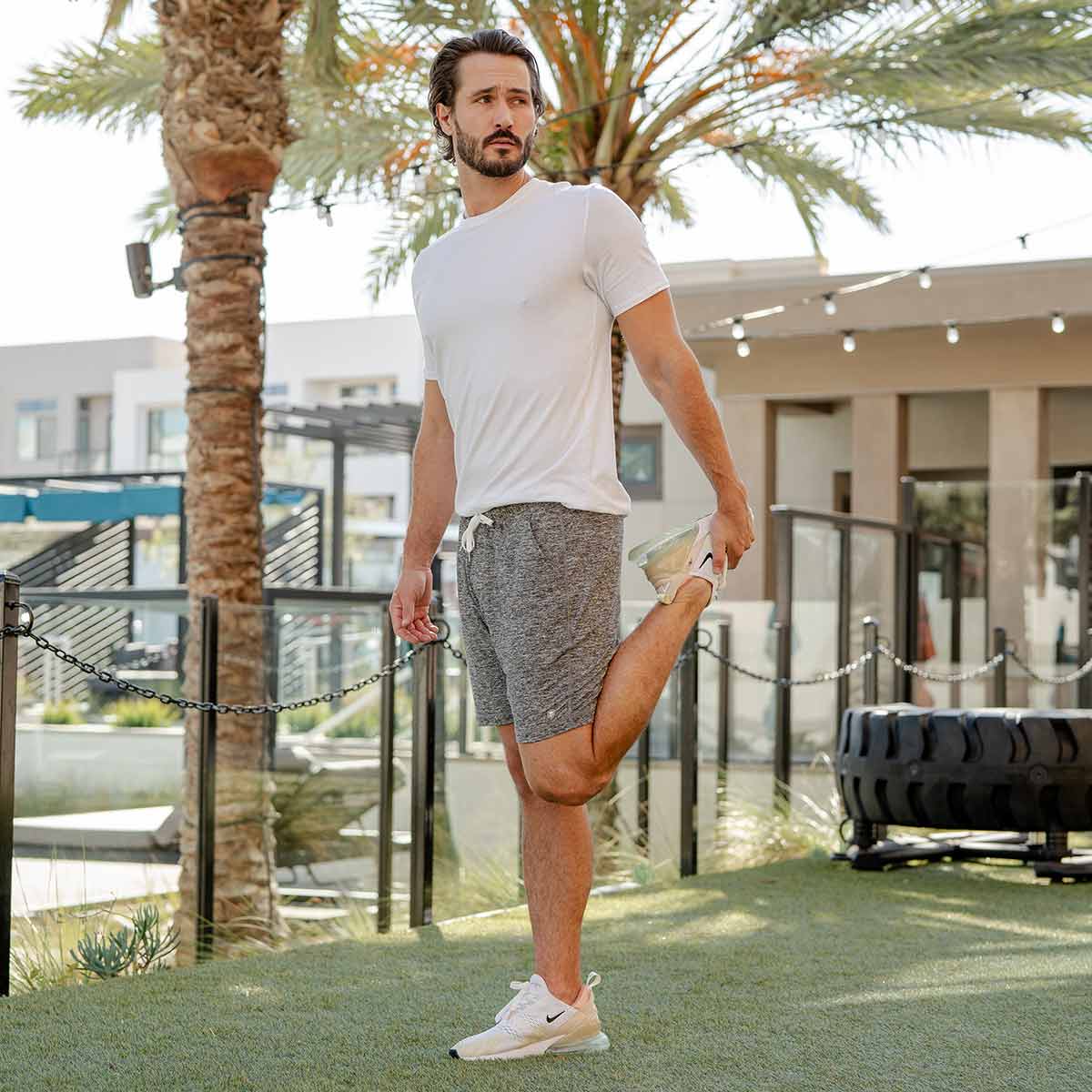 Man stretching leg outside wearing light grey Ejis Conquest athletic shorts and white tshirt