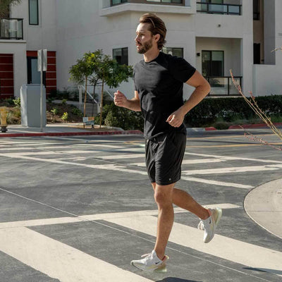 Man wearing black Ejis Conquest athletic shorts and black tshirt running on a paved path in an urban setting