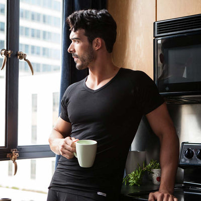 Man wearing a black Ejis v-neck micro modal sweat proof undershirt holding a green mug in a kitchen next to a window with cityscape view