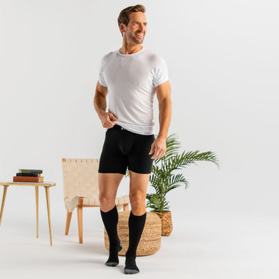 Man wearing a white Ejis crew neck micro modal sweat proof undershirt, black knee-high socks, and black sweat proof boxer briefs in a minimalistic room.