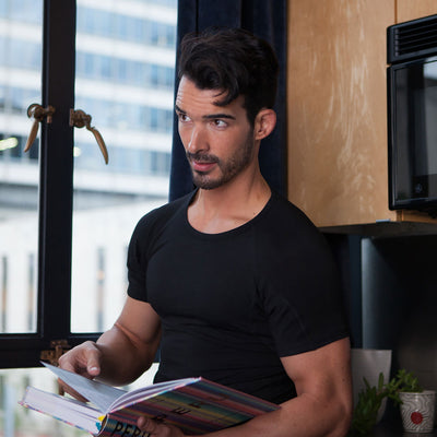 Man in a black Ejis crew neck micro modal sweat proof undershirt holding a colorful book in a kitchen.