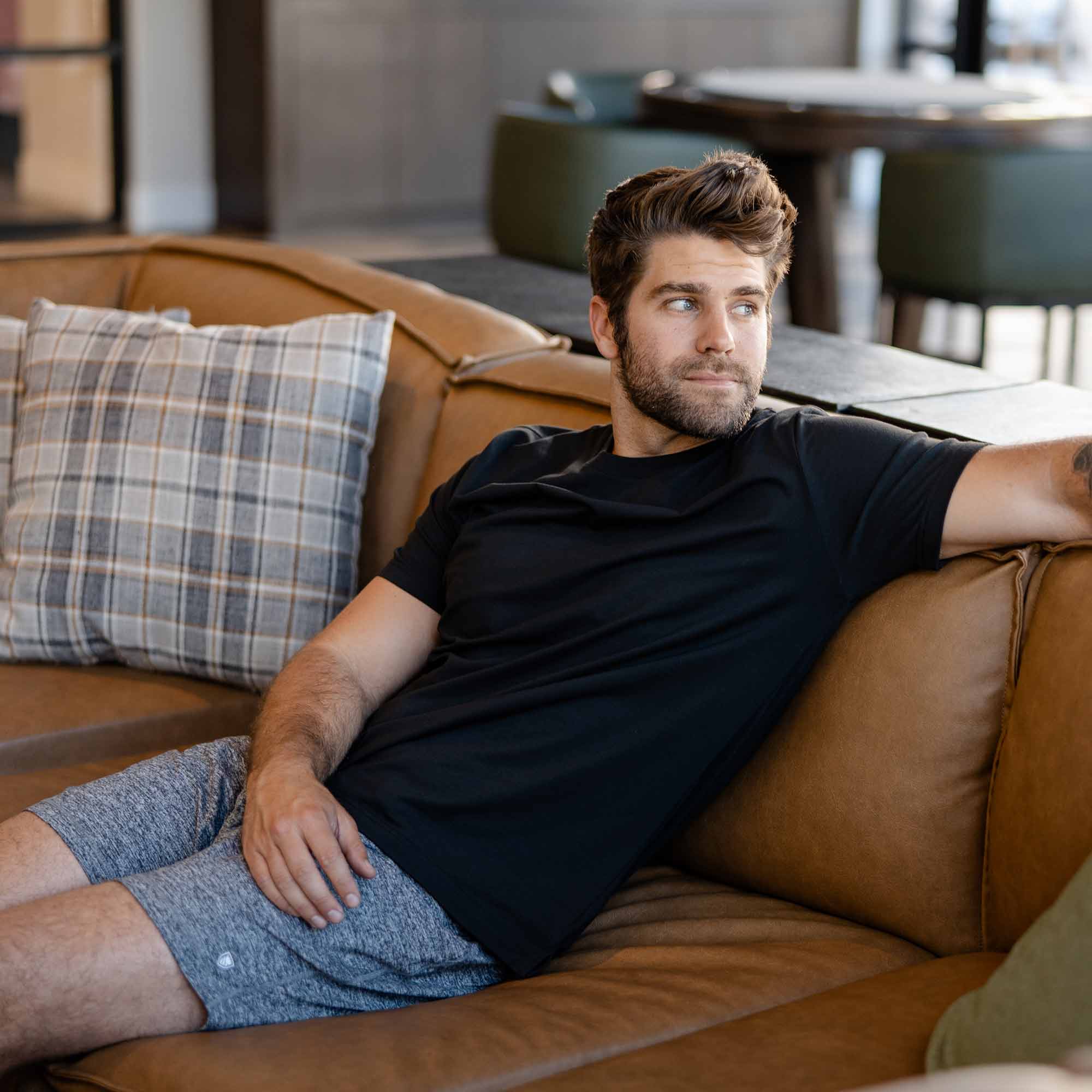 Man sitting on leather couch wearing light grey Ejis ManConquest athletic shorts and black tshirt