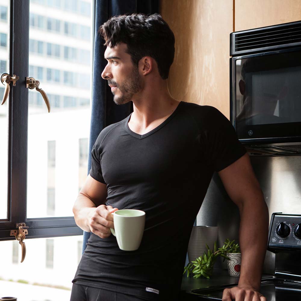 Man wearing a black Ejis v-neck micro modal sweat proof undershirt holding a green mug in a kitchen next to a window with cityscape view