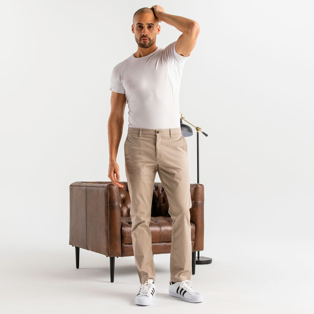 Man wearing a white Ejis crew neck micro modal sweat proof undershirt and tab pants standing next to a brown leather chair on a white background
