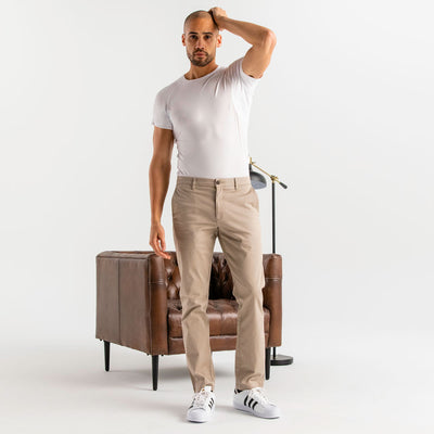 Man wearing a white Ejis crew neck micro modal sweat proof undershirt and tab pants standing next to a brown leather chair on a white background
